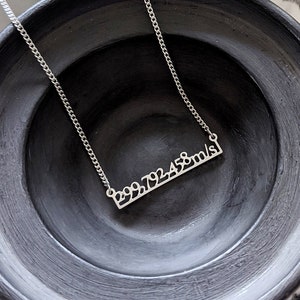 May include: A silver necklace with a bar pendant that reads "299,792,458 m/s". The necklace is displayed on a black ceramic bowl.