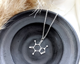 May include: A silver necklace with a pendant shaped like the chemical structure of caffeine. The necklace is displayed on a black ceramic bowl.