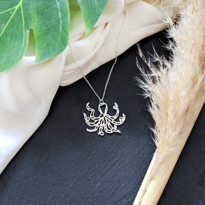 May include: A silver necklace with an intricate geometric octopus pendant. The necklace is displayed on a white fabric background with a dried grass stem.