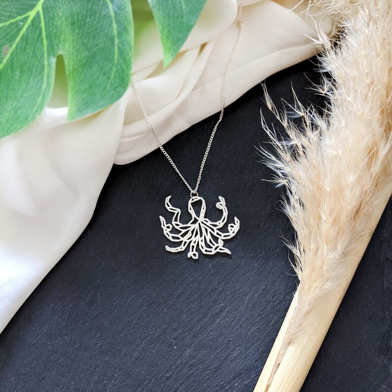 May include: A silver necklace with an intricate geometric octopus pendant. The necklace is displayed on a white fabric background with a dried grass stem.