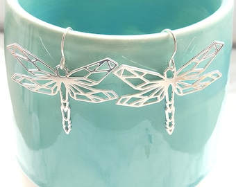 May include: A pair of silver dragonfly earrings. The earrings feature a geometric design with open wings and a detailed body. The earrings are hanging in front of a light blue ceramic mug.