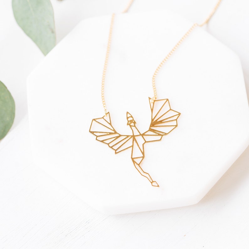 Puede incluir: Un collar de oro con un colgante de drag&oacute;n origami geom&eacute;trico. El drag&oacute;n est&aacute; en vuelo con las alas extendidas.