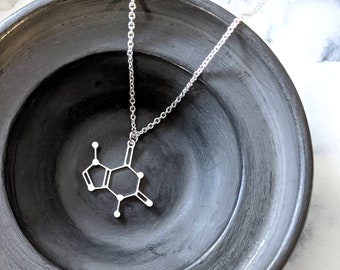 May include: A silver necklace with a pendant shaped like a chemical structure. The necklace is displayed on a black ceramic bowl.