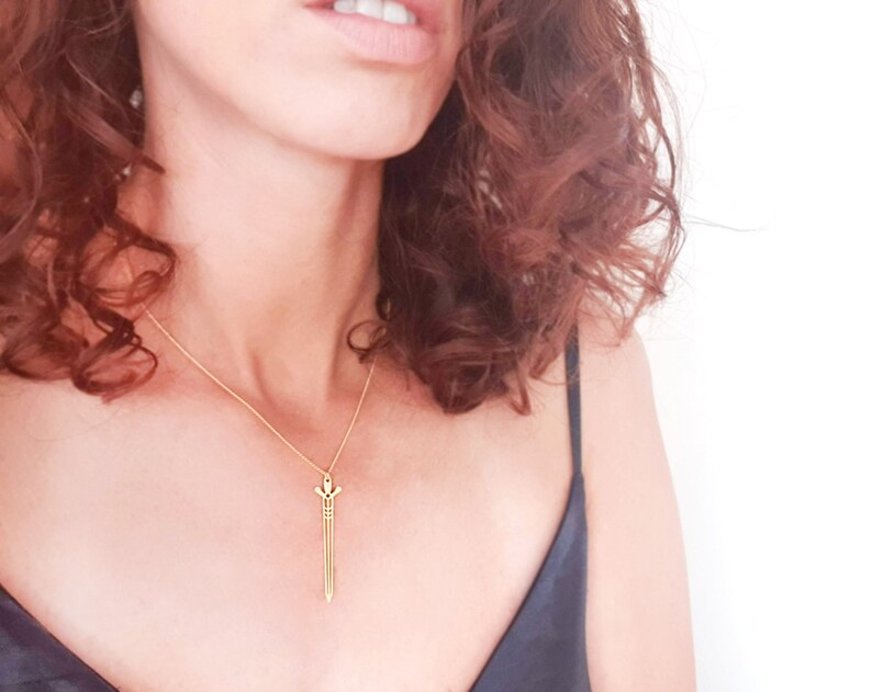 May include: A gold necklace with a sword pendant. The sword is a simple design with a pointed tip and a small cross at the top.