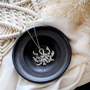 May include: A silver necklace with an intricate, geometric octopus pendant. The pendant is on a black plate, which is on a white surface.