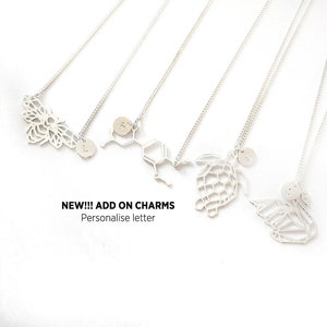 May include: Five silver necklaces with different charms, including a bee, a chemical structure, a turtle, and a butterfly. Each charm has a small circle with a letter on it. The text "NEW!!! ADD ON CHARMS Personalise letter" is below the necklaces.