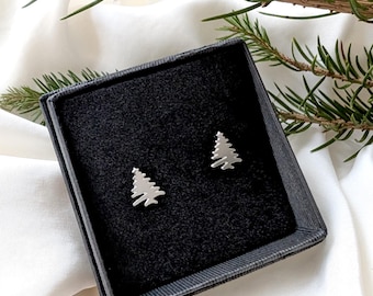 May include: A pair of silver-colored stud earrings shaped like Christmas trees, presented in a black jewelry box. The earrings are displayed against a black velvet interior, with a white fabric background and evergreen sprigs.