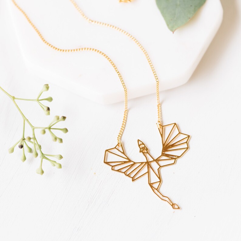 Puede incluir: Un collar de oro con un colgante de drag&oacute;n origami geom&eacute;trico. El drag&oacute;n est&aacute; en vuelo con las alas extendidas.