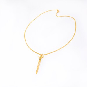 May include: A gold chain necklace with a sword pendant. The sword is a simple design with a pointed tip and a small cross at the top.