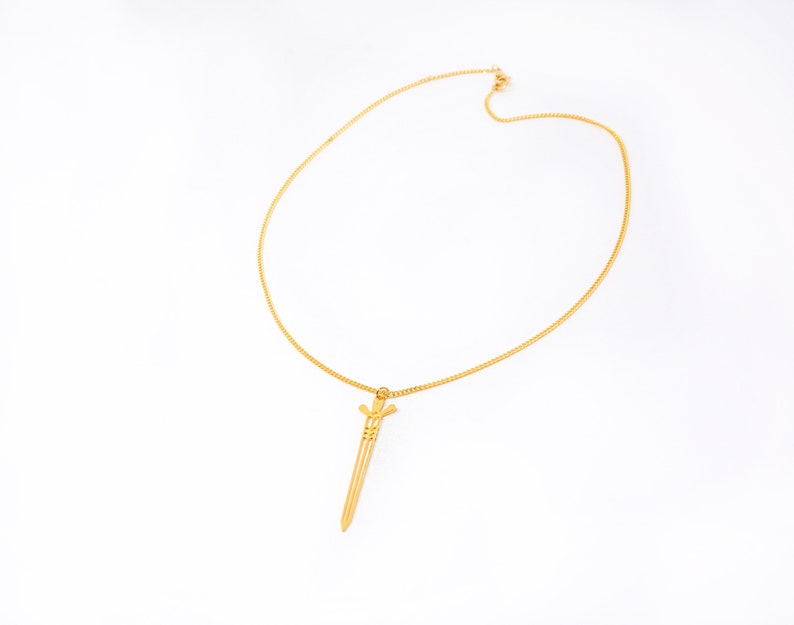 May include: A gold chain necklace with a sword pendant. The sword is a simple design with a pointed tip and a small cross at the top.