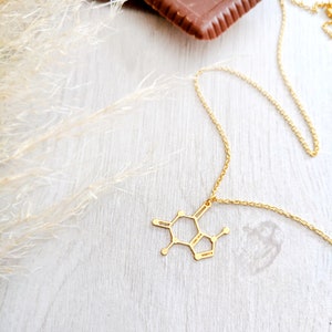 May include: A gold necklace with a caffeine molecule pendant. The necklace is on a white wooden surface with a piece of chocolate in the background.