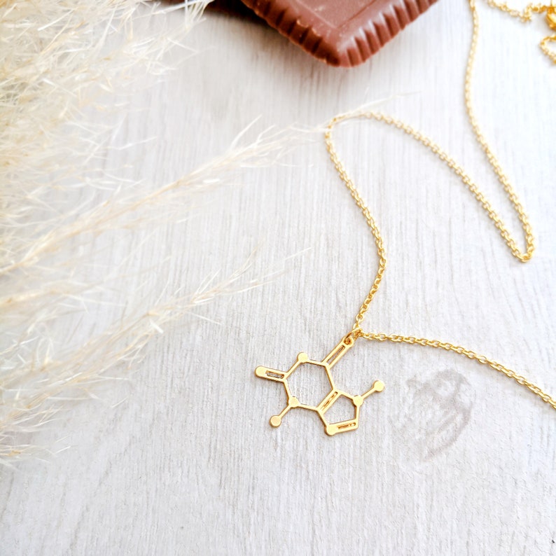May include: A gold necklace with a caffeine molecule pendant. The necklace is on a white wooden surface with a piece of chocolate in the background.