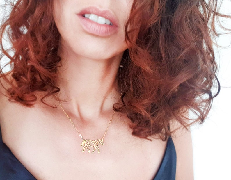 May include: A gold necklace featuring a delicate origami horse pendant. The necklace is worn by a person with long, curly red hair.