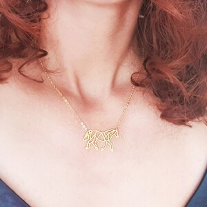 May include: A gold necklace featuring a geometric horse pendant. The necklace is worn by a person with long, curly red hair.