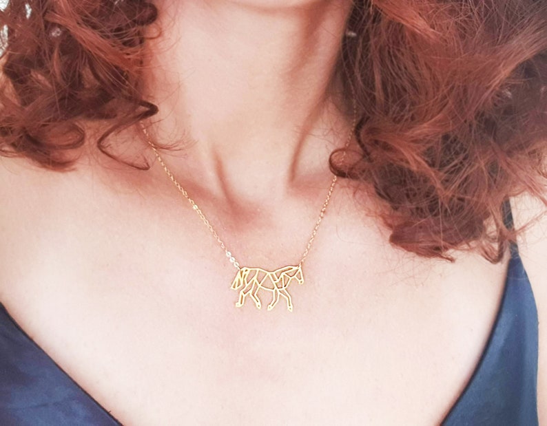 May include: A gold necklace featuring a geometric horse pendant. The necklace is worn by a person with long, curly red hair.