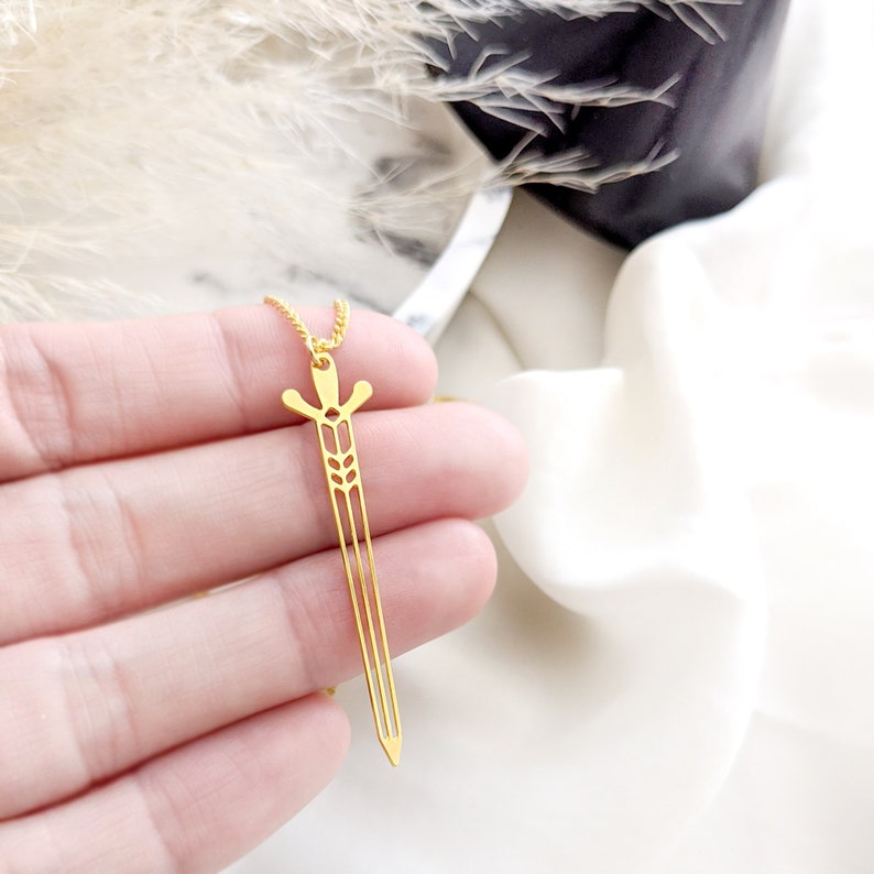 May include: A gold-toned necklace with a sword pendant. The sword is a cutout design with a wheat stalk pattern.