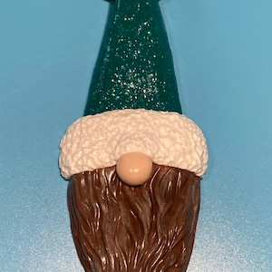 Peut inclure: Une décoration de gnome verte et blanche avec une barbe brune. Le gnome porte un chapeau vert avec une bordure blanche et un pompon blanc sur le dessus. Le gnome est suspendu à un cordon noir.