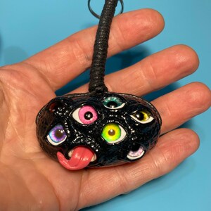May include: Black leather keychain featuring a 3D eye monster design. The monster has a pink tongue sticking out and multiple colourful eyes.