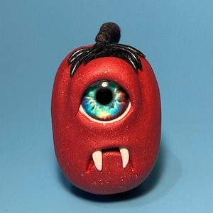 May include: A red, one-eyed monster with fangs and black hair. The monster has a blue and green eye with a white pupil.