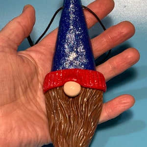 Peut inclure: Une décoration de gnome bleue et rouge avec un chapeau bleu pailleté et une barbe brune. Le gnome est suspendu à une ficelle noire.