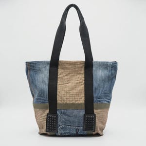 Op de afbeelding: Een tote bag gemaakt van een patchwork van blauwe denim en beige stof met een rasterachtig stikselpatroon. De tas heeft twee zwarte stoffen hengsels en twee zwarte rechthoekige patches aan de onderkant, elk versierd met kleine zwarte studs.