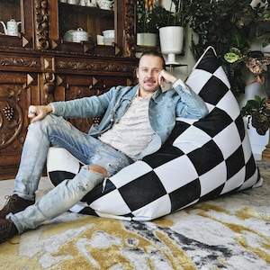 May include: A person sits in a black and white checkered bean bag chair. The chair is made of fabric and has a square pattern. The person is wearing blue jeans and a denim jacket.