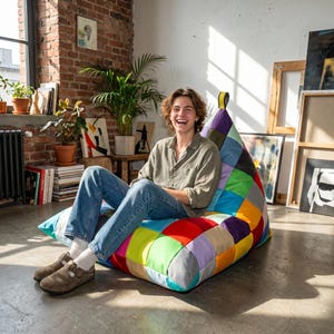 Könnte beinhalten: Ein dreieckiger Sitzsack mit einem lebendigen Patchwork-Design, das aus zahlreichen quadratischen Stoffbahnen in verschiedenen Farben wie Rot, Blau, Grün, Gelb, Lila und Orange besteht, mit einer kleinen gelben Stoffschlaufe an der Spitze.