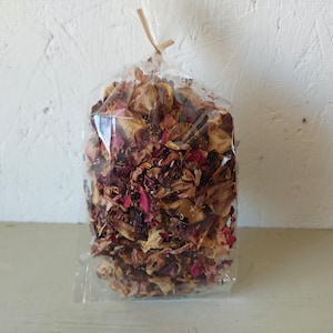 May include: A clear plastic bag filled with dried rose petals. The petals are a mix of brown, beige, and pink hues. The bag is tied at the top with a natural twine. The background is a textured white wall.