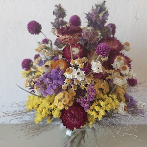 May include: A bouquet of dried flowers featuring deep purple globe amaranth, vibrant yellow statice, white strawflowers, and dark red blossoms. The arrangement includes various textures of dried stems and foliage, creating a dense, colorful cluster.