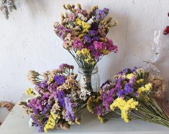 May include: A collection of dried statice flowers in vibrant shades of purple, magenta, yellow, and cream. One bouquet is arranged upright in a clear glass mason jar, while two additional bouquets are laid horizontally on a flat surface.