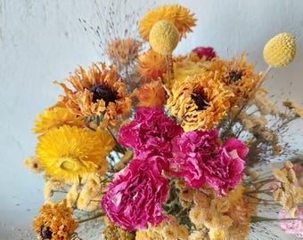 May include: A close-up of a dried flower arrangement. The bouquet features a variety of textures and colours, including bright yellow, orange, and pink flowers, with delicate seed heads and stems. The background is a neutral colour.