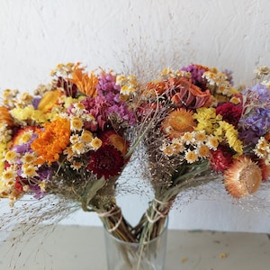 May include: A bouquet of dried flowers featuring a vibrant mix of orange, yellow, purple, and deep red blossoms. The arrangement includes various small-petaled flowers and wispy grasses, all tied together with twine and displayed in a clear glass vase.