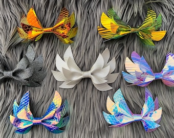 Könnte beinhalten: Acht verschiedene Haarbänder mit verschiedenen schillernden und glitzernden Oberflächen. Die Schleifen sind flügelartig geformt und in zwei Reihen auf einer grauen, flauschigen Oberfläche angeordnet.
