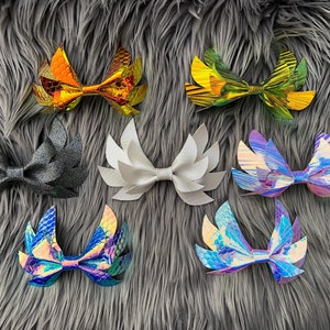 May include: Eight different hair bows with various iridescent and glitter finishes. The bows are shaped like wings and are arranged in two rows on a gray fuzzy surface.