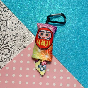 May include: A Daruma doll keychain with a black carabiner clip. The doll is red with a white face and the Japanese character for "fortune". The fabric has a colorful geometric pattern. A small fabric tag with a floral design is attached.
