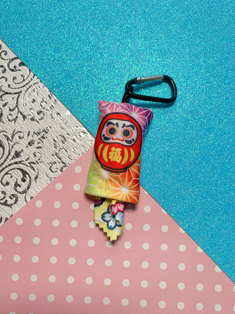 May include: A Daruma doll keychain with a black carabiner clip. The doll is red with a white face and the Japanese character for "fortune". The fabric has a colorful geometric pattern. A small fabric tag with a floral design is attached.