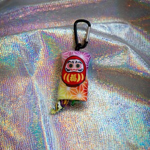 May include: A colorful Daruma doll keychain with a black carabiner clip. The Daruma doll has a red face, a yellow body, and the Japanese character for "fortune" in the center. The background is a shiny, iridescent fabric.
