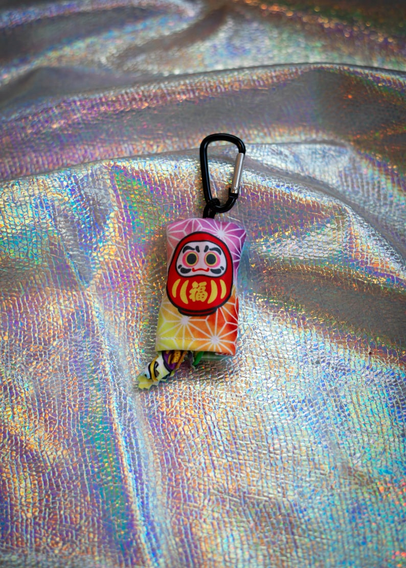 May include: A colorful Daruma doll keychain with a black carabiner clip. The Daruma doll has a red face, a yellow body, and the Japanese character for "fortune" in the center. The background is a shiny, iridescent fabric.