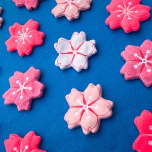 May include: A collection of pink and white felt cherry blossom flowers on a blue background. The flowers are all different sizes and have a simple, embroidered design.