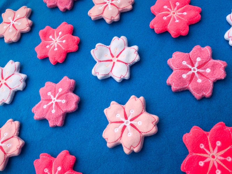 May include: A collection of pink and white felt cherry blossom flowers on a blue background. The flowers are all different sizes and have a simple, embroidered design.