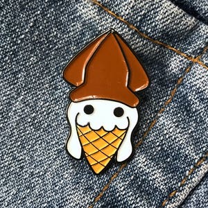 Op de afbeelding: Een bruine en witte emaille pin van een inktvis die een ijshoornhoed draagt. De inktvis heeft een wit lichaam en bruine ogen. De ijshoorn is geel en oranje met een witte swirl van ijs.