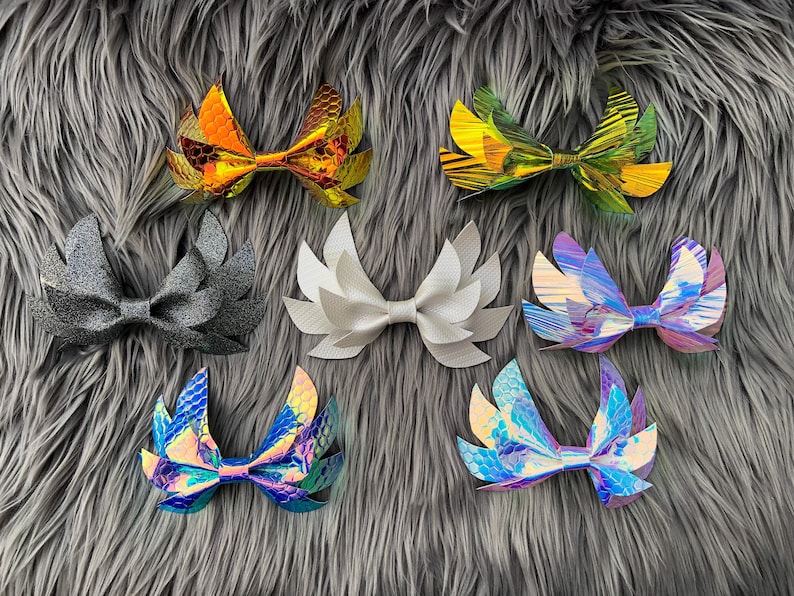May include: Eight different hair bows with various iridescent and glitter finishes. The bows are shaped like wings and are arranged in two rows on a gray fuzzy surface.