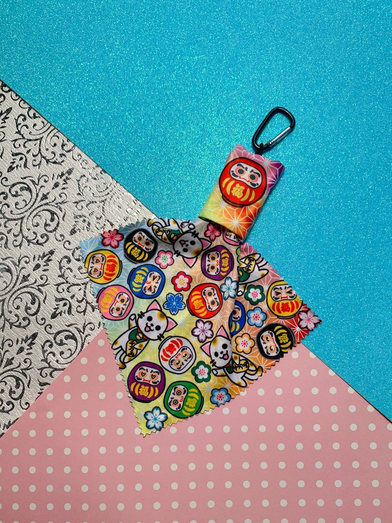 May include: A Daruma doll keychain with a black carabiner clip. The keychain has a colorful gradient design and a Daruma doll face. Two patterned cloths feature Daruma dolls and cartoon cats with flowers. The background is a mix of pink, blue, and white.