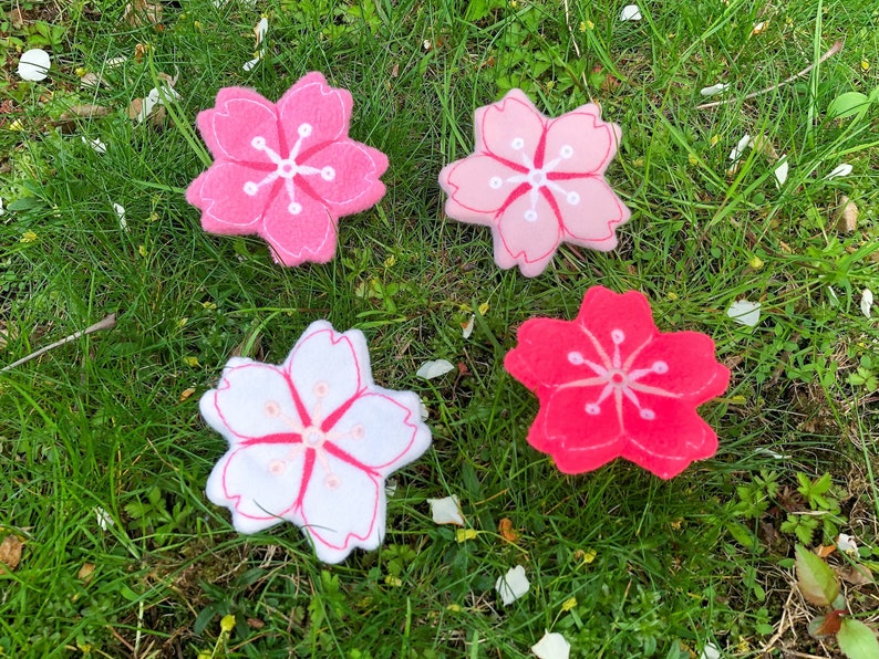 May include: Four felt cherry blossom flowers in pink, white, and red shades. The flowers are arranged on a bed of green grass.