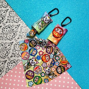 May include: Two small, colorful pouches with black carabiner clips, featuring Japanese Daruma doll and cat designs. A matching fabric with similar patterns is also displayed. The pouches have a gradient color scheme, with a variety of colors.