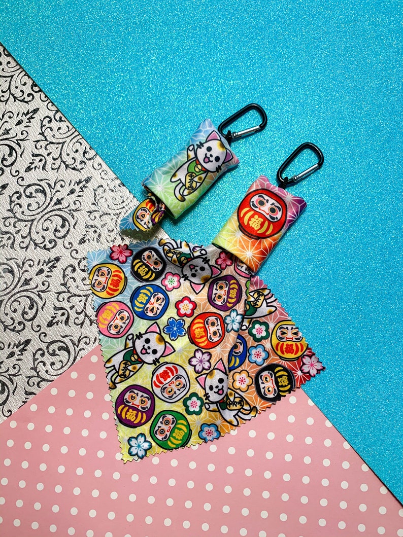 May include: Two small, colorful pouches with black carabiner clips, featuring Japanese Daruma doll and cat designs. A matching fabric with similar patterns is also displayed. The pouches have a gradient color scheme, with a variety of colors.