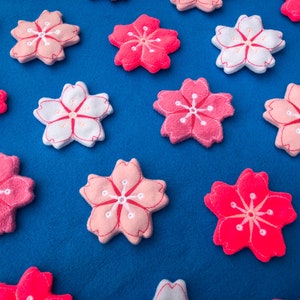 May include: A collection of felt cherry blossom flowers in pink and white with embroidered details. The flowers are arranged on a blue background.