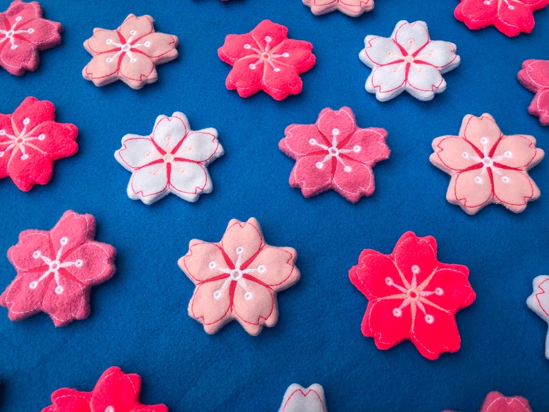 May include: A collection of felt cherry blossom flowers in pink and white with embroidered details. The flowers are arranged on a blue background.