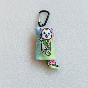 May include: A colorful Maneki-neko cat charm with a black carabiner clip. The charm features a smiling cat with a gold coin, set against a geometric pattern. The cat's tail has a floral design.