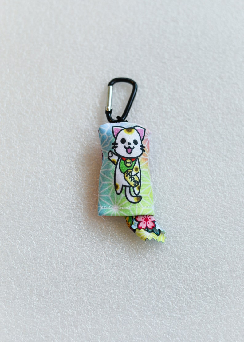 May include: A colorful Maneki-neko cat charm with a black carabiner clip. The charm features a smiling cat with a gold coin, set against a geometric pattern. The cat's tail has a floral design.